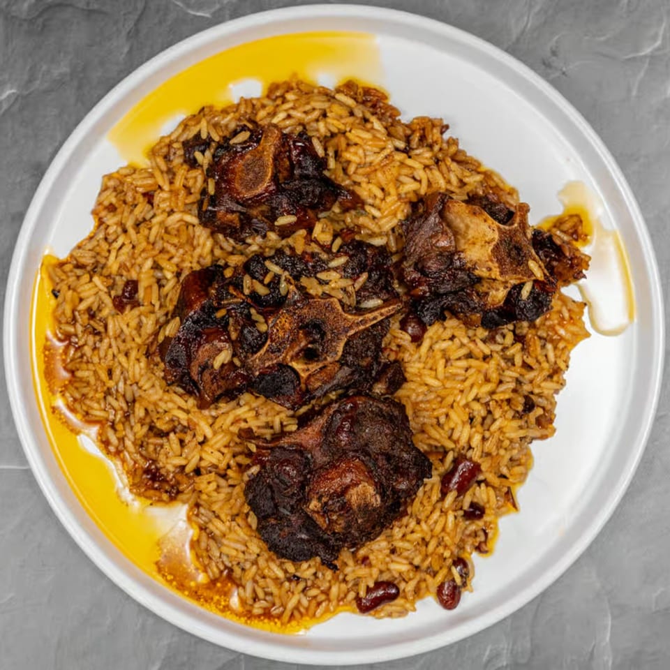 A white plate holds a serving of Jamaican oxtail stew with rice and peas, glistening with sauce.