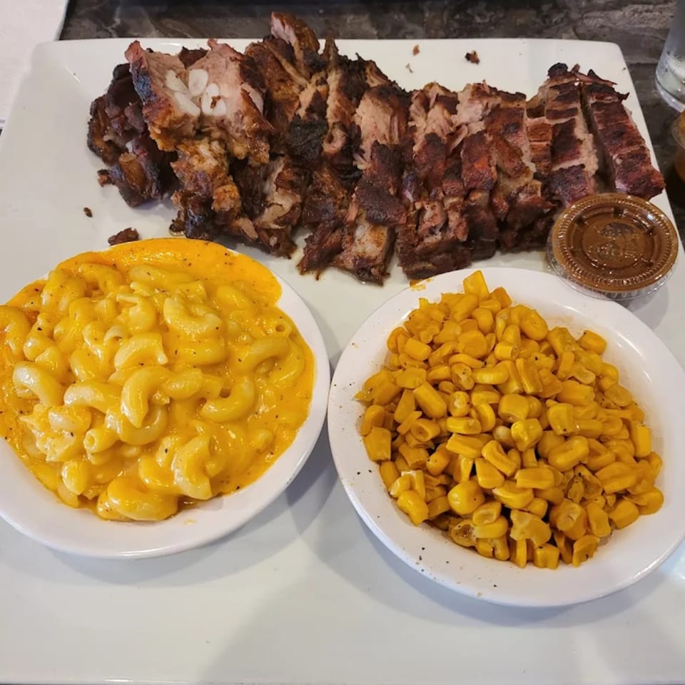 Jerk pork with macaroni and cheese, corn, and a side of sauce on a white plate.