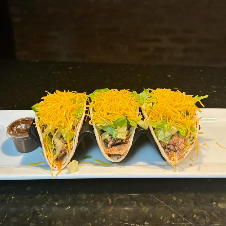 Three Jerk Villa tacos filled with meat, lettuce, and shredded cheese, served with a side of sauce.