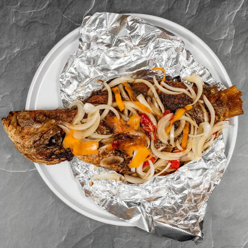 Whole fried fish, topped with sautéed onions and bell peppers, served on foil on a white plate.